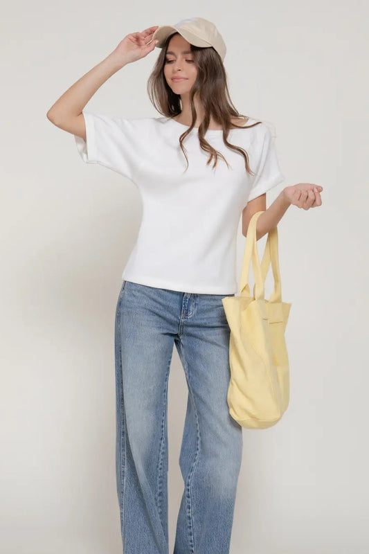 Woman wearing a white shirt, blue jeans, and a beige cap, holding a yellow tote bag against a plain background.