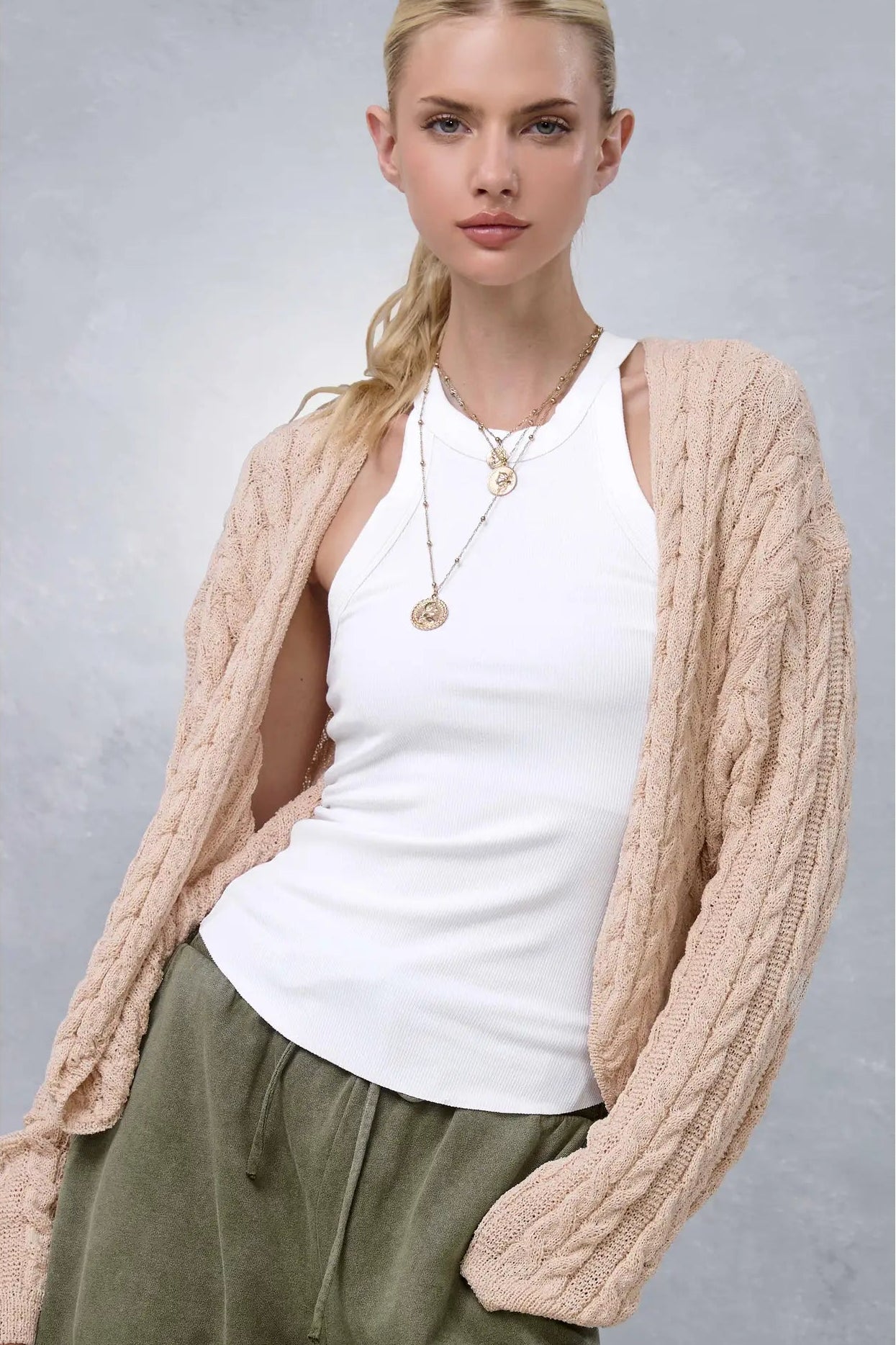 A woman wearing a beige cable-knit cardigan over a white tank top and olive green pants, standing against a textured gray studio background.