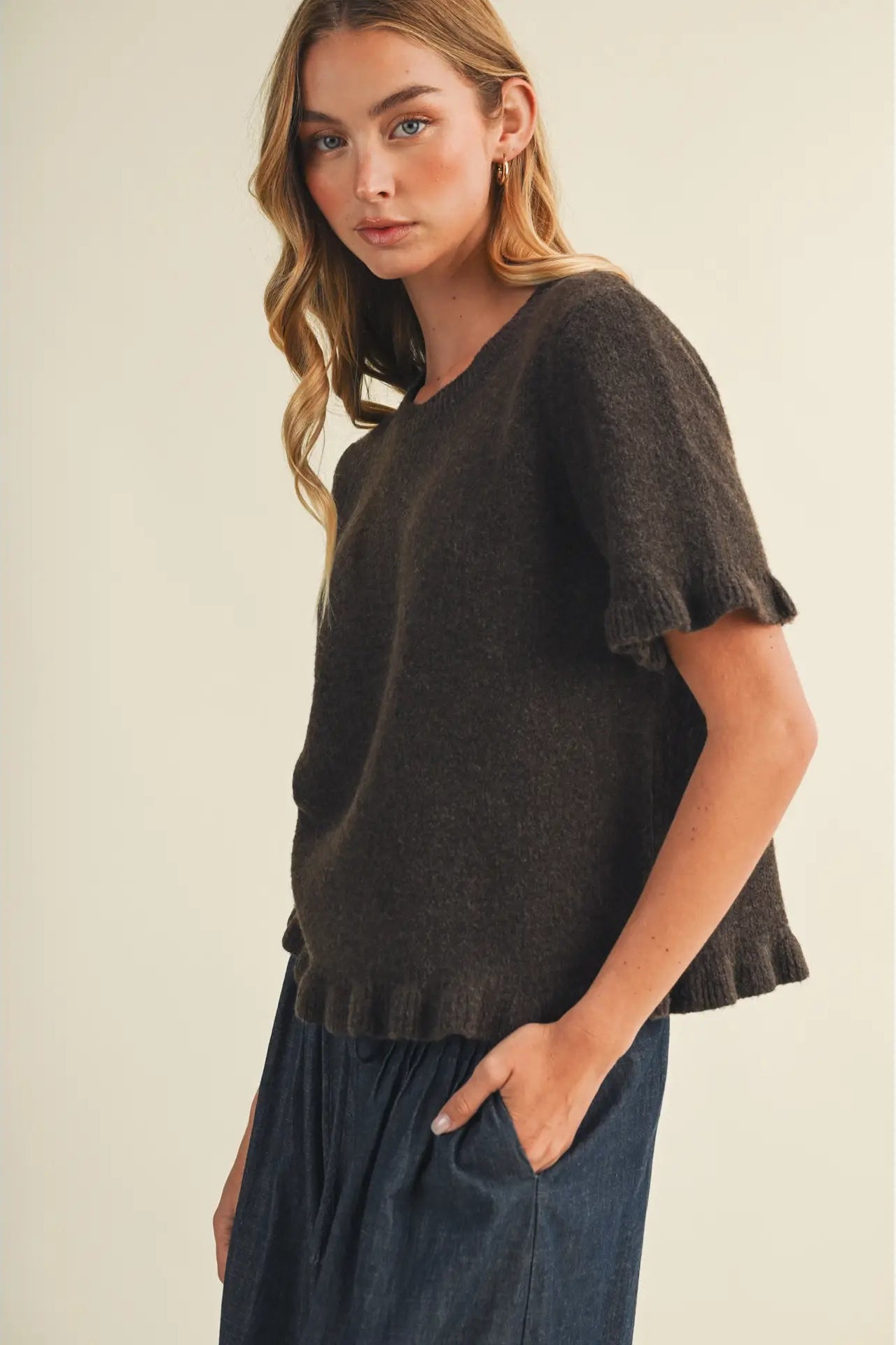A woman wearing a dark brown short-sleeve knit top with ruffled edges and dark blue pants, standing against a plain beige studio background.