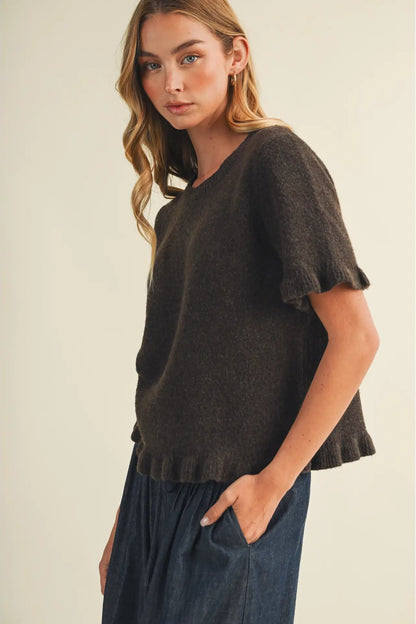 A woman wearing a dark brown short-sleeve knit top with ruffled edges and dark blue pants, standing against a plain beige studio background.