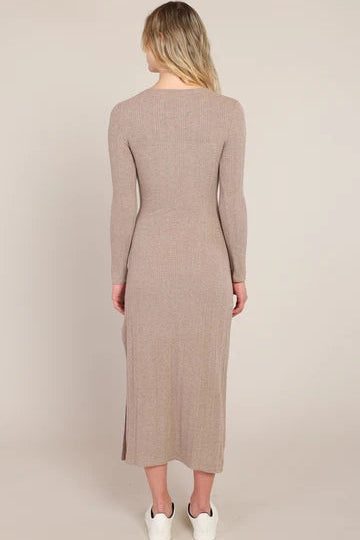 A woman viewed from the back wearing a long-sleeve, knee-length knit dress in a light taupe-beige color, paired with white sneakers, against a solid light beige studio background.