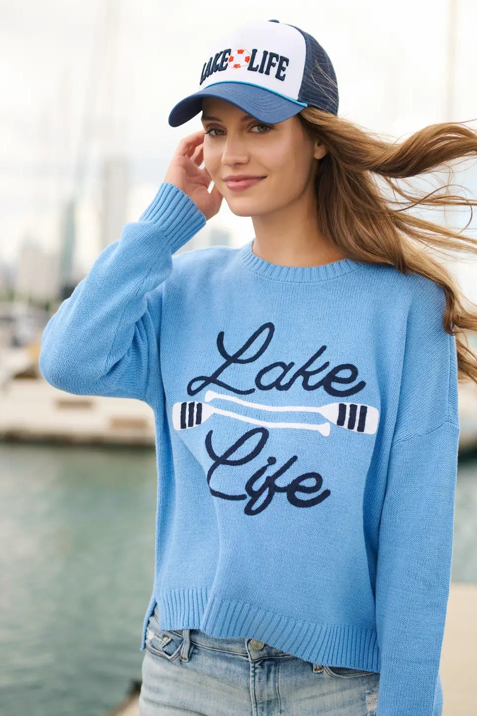 Woman wearing a blue 'Lake Life' sweater and cap by a waterfront.