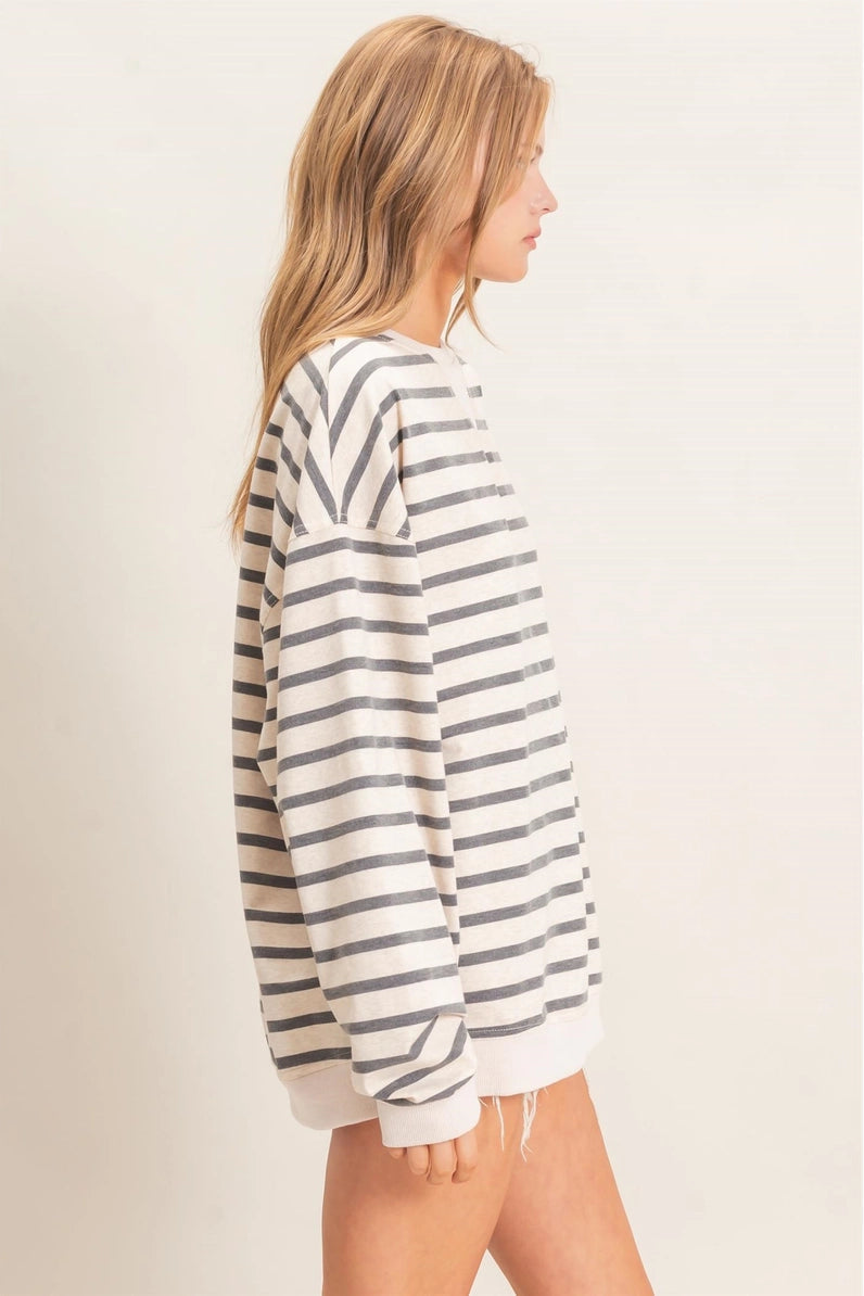 A woman shown from the side wearing an oversized long-sleeve knit top with horizontal gray and off-white stripes, photographed against a plain light beige studio background.