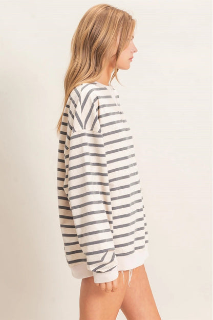 A woman shown from the side wearing an oversized long-sleeve knit top with horizontal gray and off-white stripes, photographed against a plain light beige studio background.