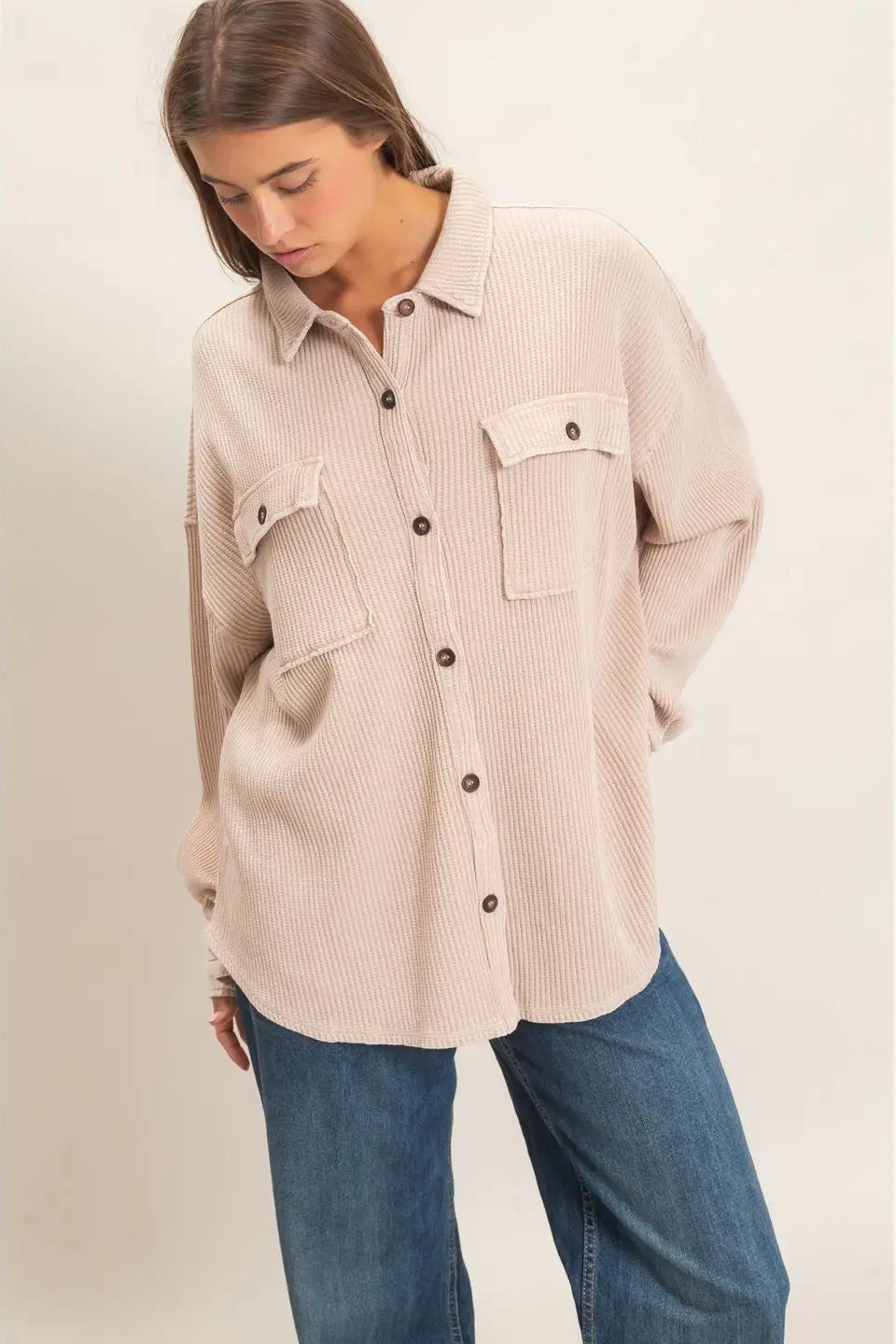 A woman wearing a light beige ribbed button-up shirt with chest pockets and blue jeans, standing against a plain light beige studio background.