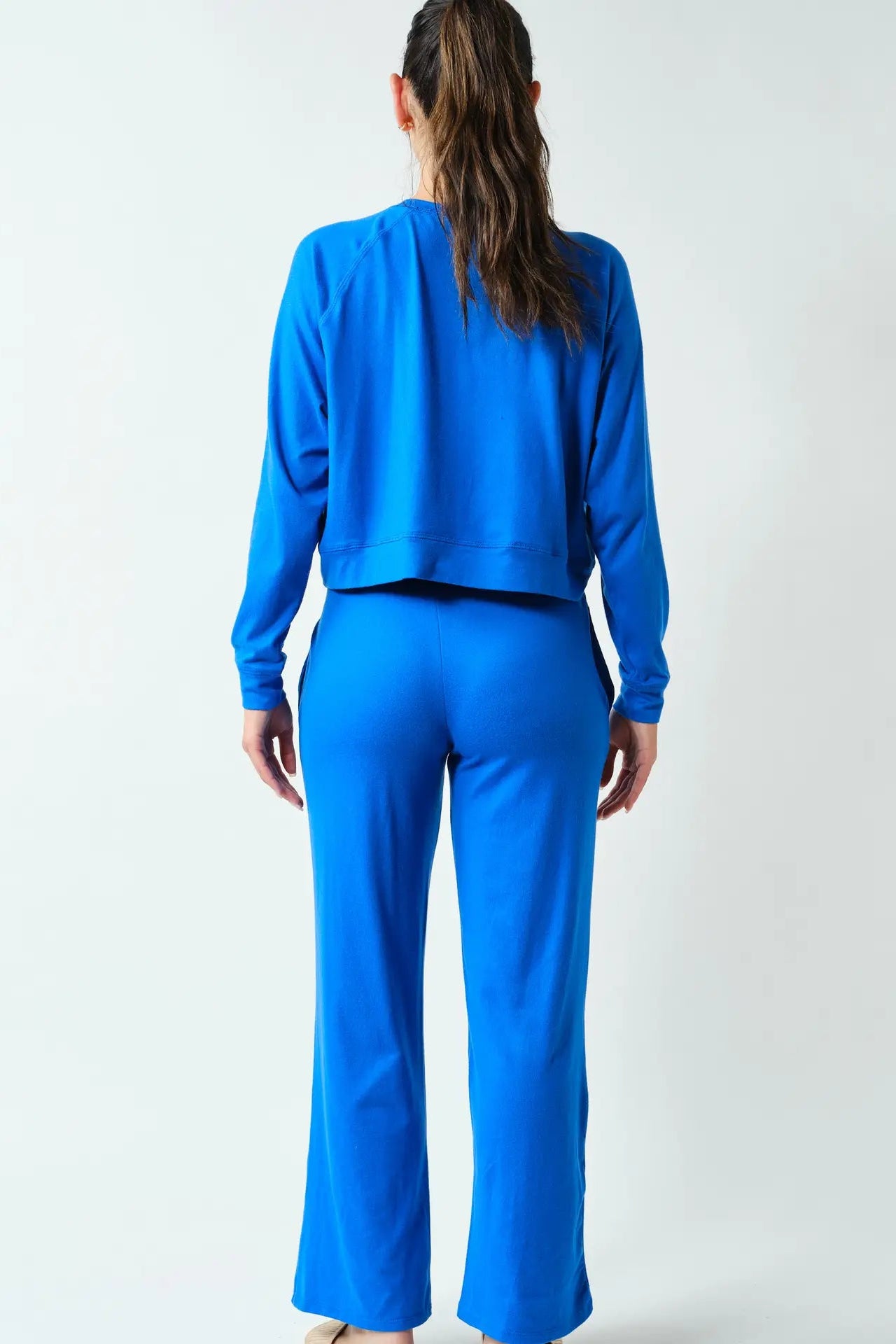 A woman viewed from behind wearing a bright blue matching lounge set consisting of a cropped long-sleeve top and wide-leg pants, against a plain light gray studio background.