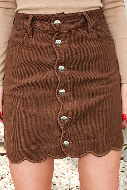 A brown corduroy mini skirt with a scalloped front edge and silver-tone button closures, shown on a model wearing a beige long-sleeve top.