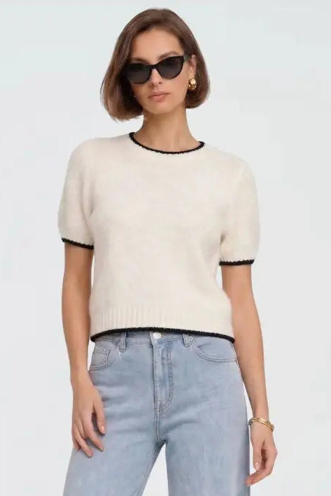 A woman modeling a short-sleeve cream knit sweater with black trim on the crew neck and sleeve cuffs, paired with light blue jeans against a plain light gray background.