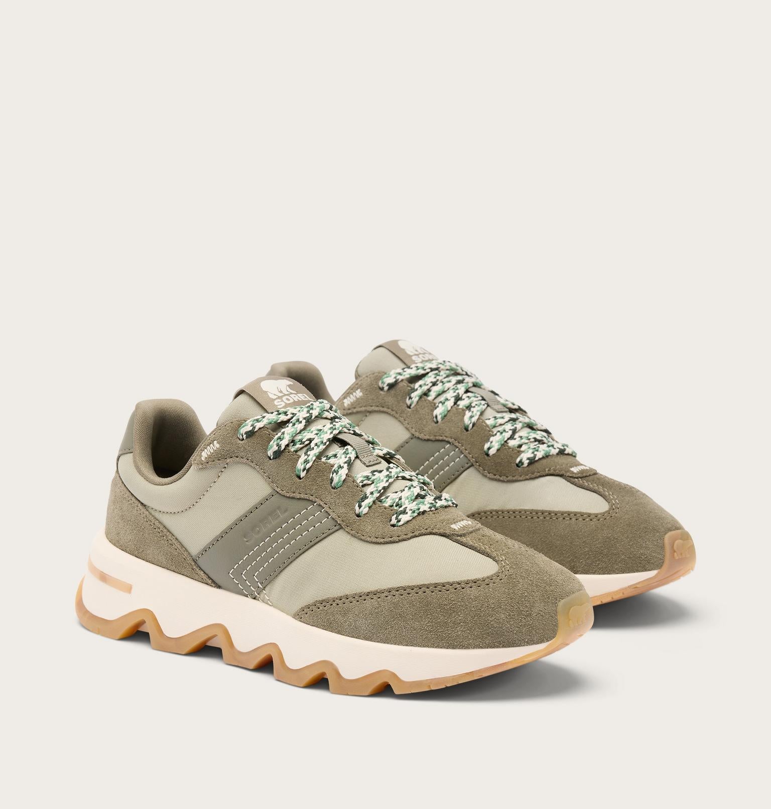 A pair of low-top sneakers in muted olive green and beige suede and mesh, featuring chunky cream midsoles and patterned green-and-white laces, photographed on a light beige studio background.