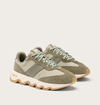 A pair of low-top sneakers in muted olive green and beige suede and mesh, featuring chunky cream midsoles and patterned green-and-white laces, photographed on a light beige studio background.