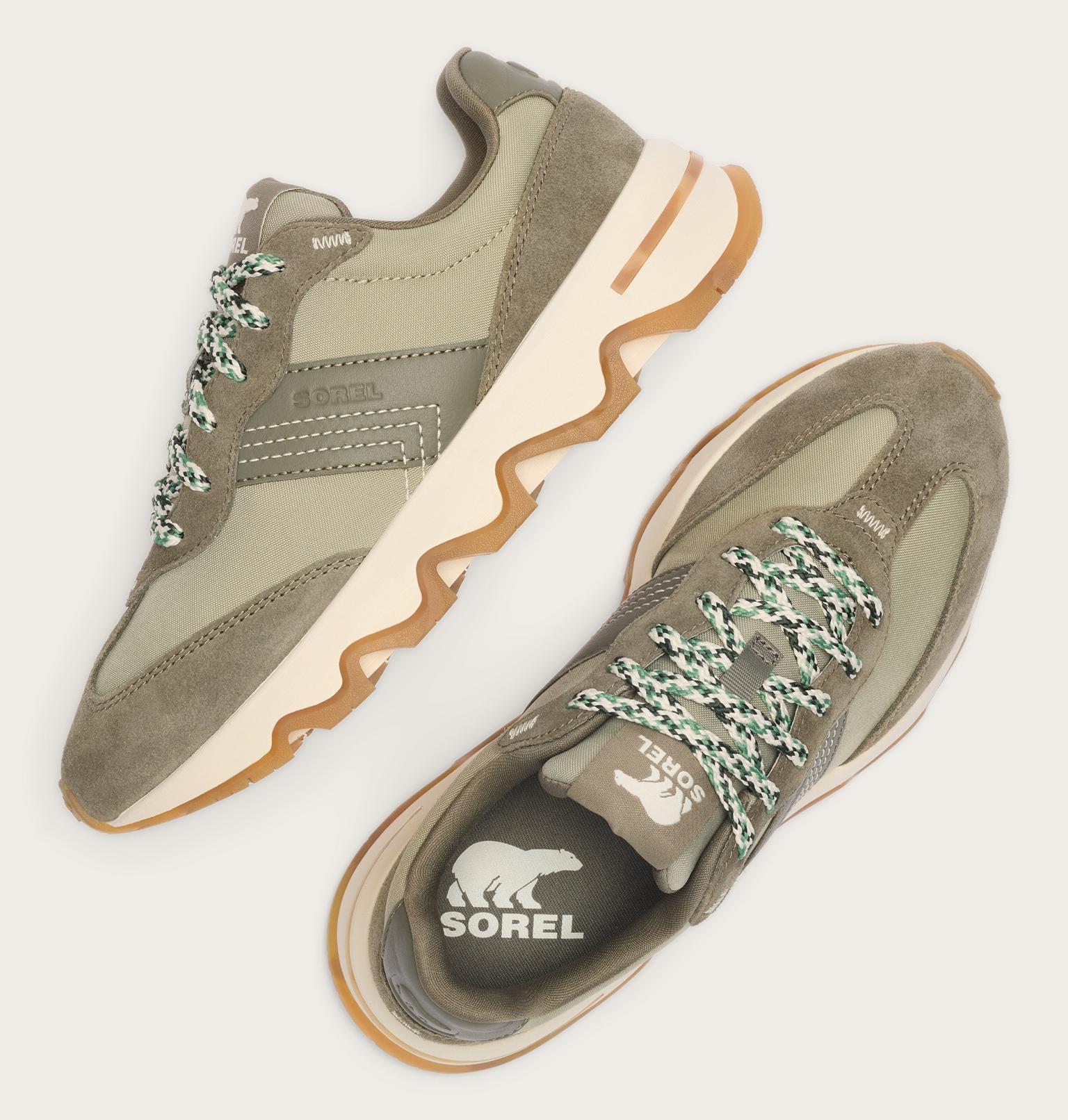 A pair of Sorel sneakers in olive green suede and mesh, featuring chunky wavy beige midsoles, gum brown outsoles, and green-and-white patterned laces, shown from top-down angles on a light beige background.