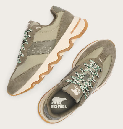 A pair of Sorel sneakers in olive green suede and mesh, featuring chunky wavy beige midsoles, gum brown outsoles, and green-and-white patterned laces, shown from top-down angles on a light beige background.