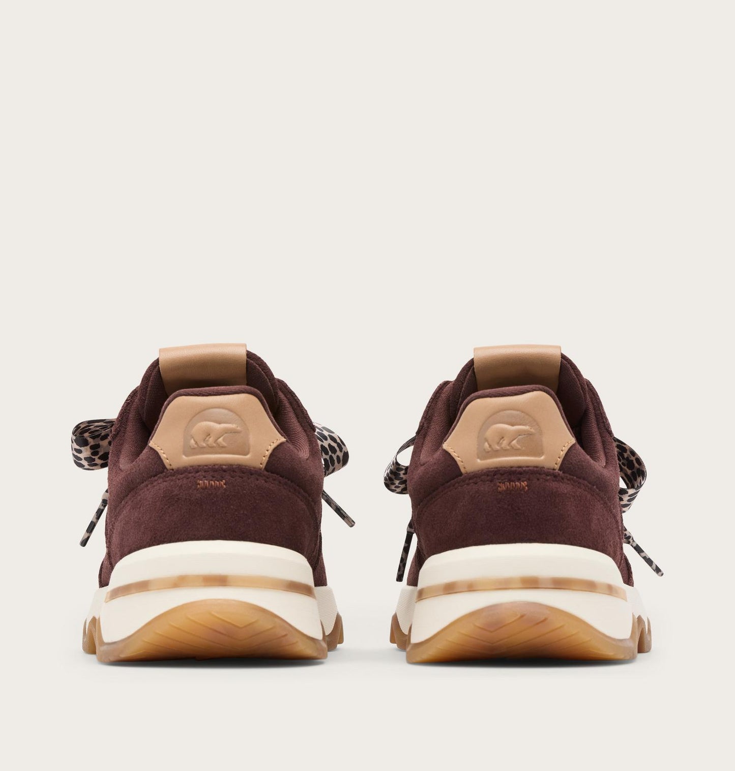 A pair of low-top sneakers shown from the back, featuring dark burgundy suede and tan leather panels, a thick cream midsole, and patterned laces, set against a solid off-white background.