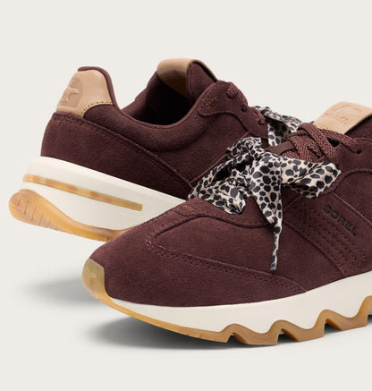 A pair of Sorel sneakers in deep burgundy suede with beige leather accents, featuring a patterned leopard-print ribbon tied around the laces, set against a light gray background.