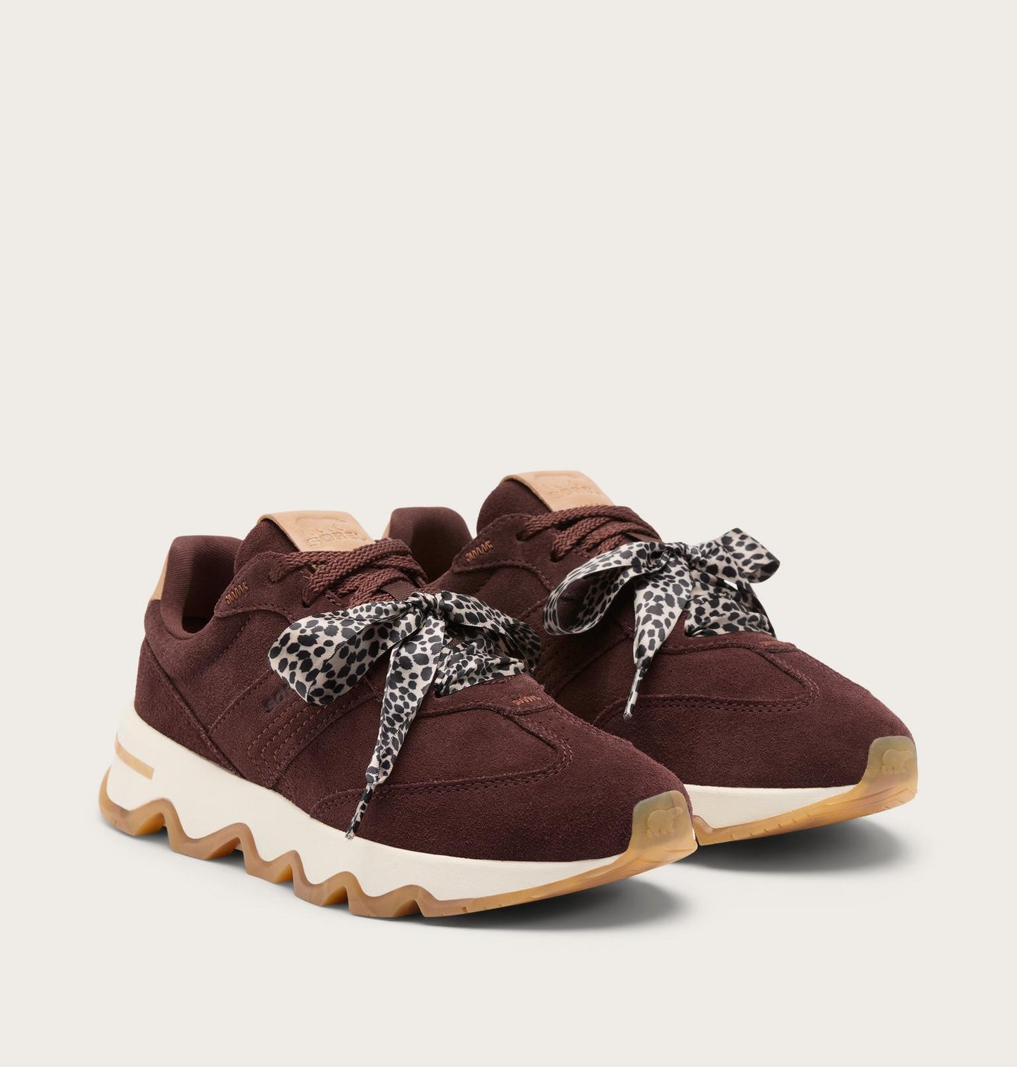 A pair of low-top sneakers in dark burgundy suede with patterned leopard-print laces and chunky wavy white and gum soles, photographed on a light beige background.