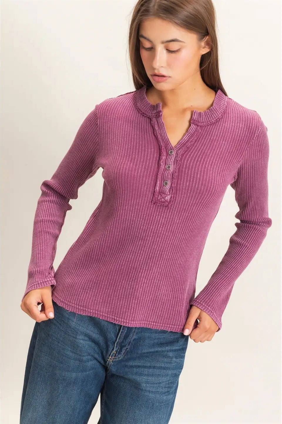 A woman wearing a ribbed long-sleeve henley top in dusty mauve pink with blue jeans, standing against a plain light beige studio background.