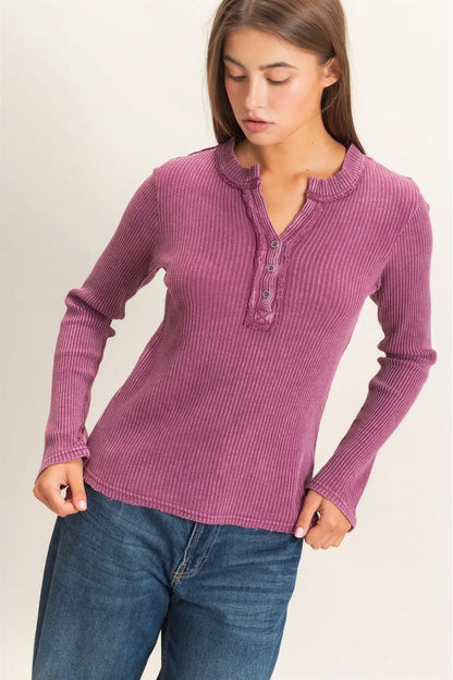 A woman wearing a ribbed long-sleeve henley top in dusty mauve pink with blue jeans, standing against a plain light beige studio background.