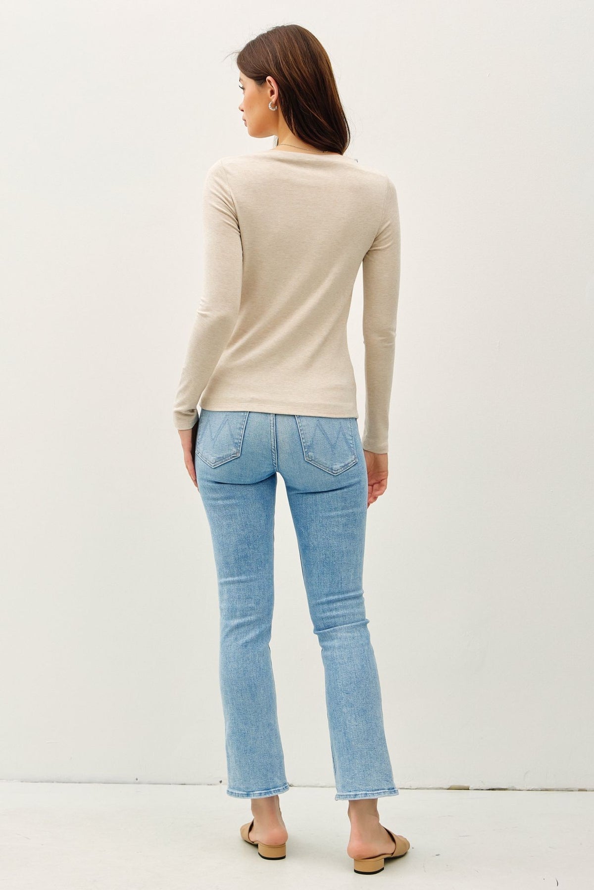 A woman shown from the back wearing a beige long-sleeve knit top and light blue cropped flare jeans, standing against a plain off-white wall.