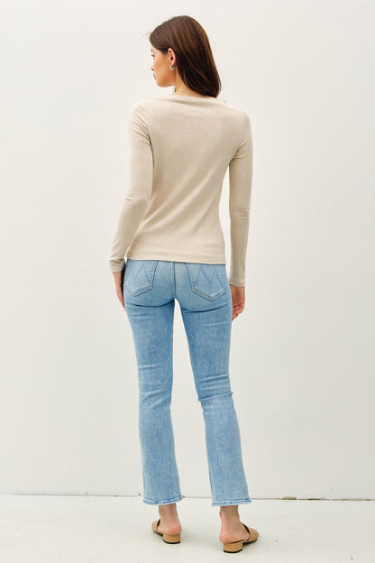 A woman shown from the back wearing a beige long-sleeve knit top and light blue cropped flare jeans, standing against a plain off-white wall.