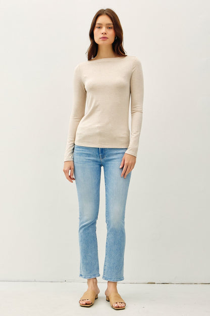 A woman modeling a beige long-sleeve knit top with a boat neckline, paired with light-wash blue jeans and tan sandals, standing against a plain white studio background.