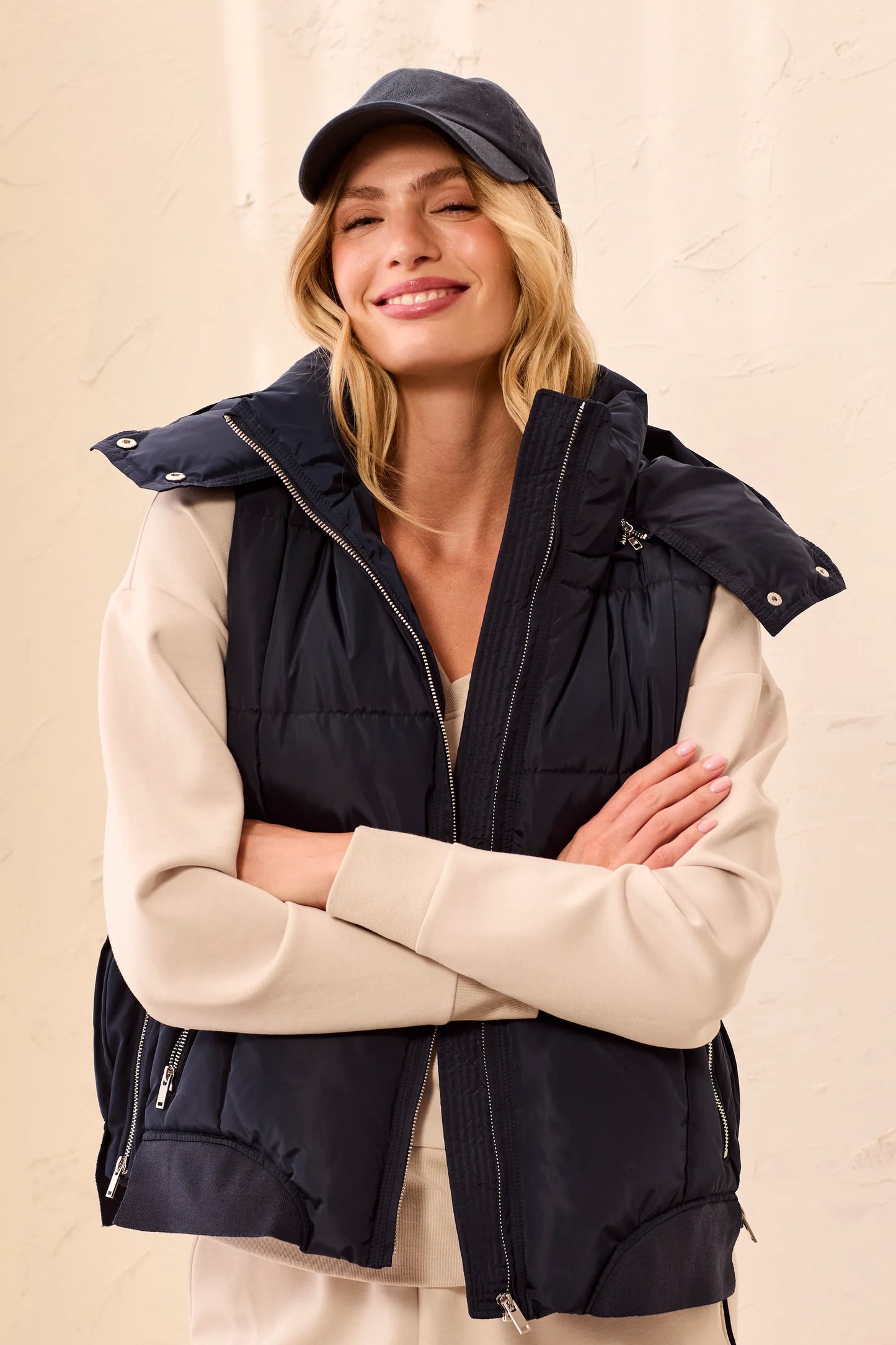 A woman wearing a navy blue puffer vest over a beige long-sleeve top and matching pants, accessorized with a dark baseball cap, posing with her arms crossed against a textured beige wall.