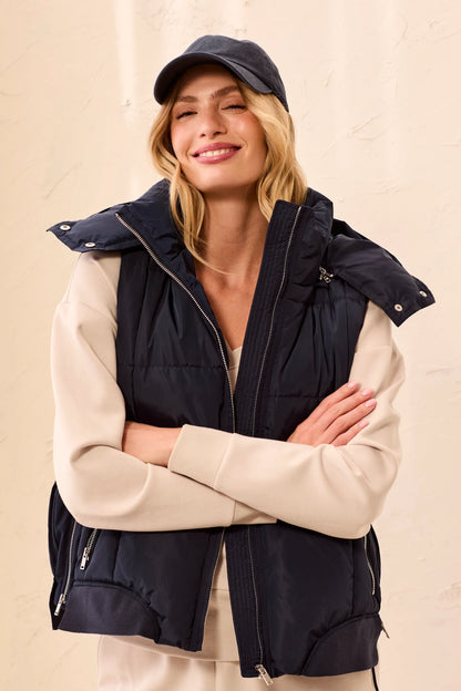 A woman wearing a navy blue puffer vest over a beige long-sleeve top and matching pants, accessorized with a dark baseball cap, posing with her arms crossed against a textured beige wall.
