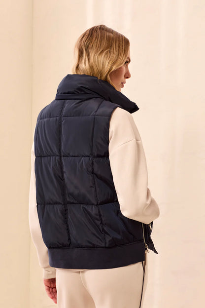 A woman shown from the back wearing a dark navy puffer vest over a cream-colored long-sleeve top and cream pants, set against a soft beige background.