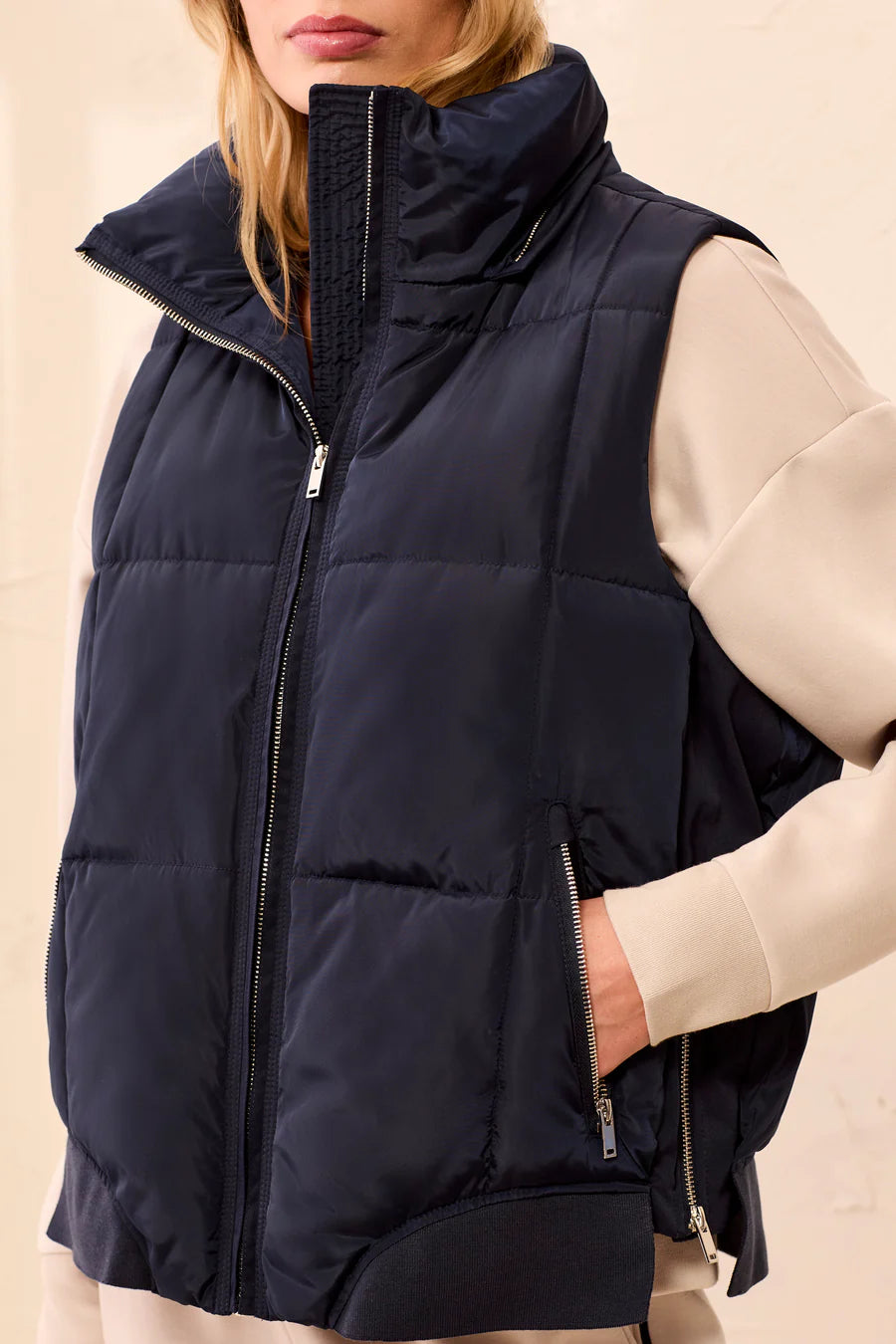 A close-up of a model wearing a navy blue puffer vest over a cream-colored long-sleeve top. The vest features a high collar, a front zipper, and zippered side pockets.