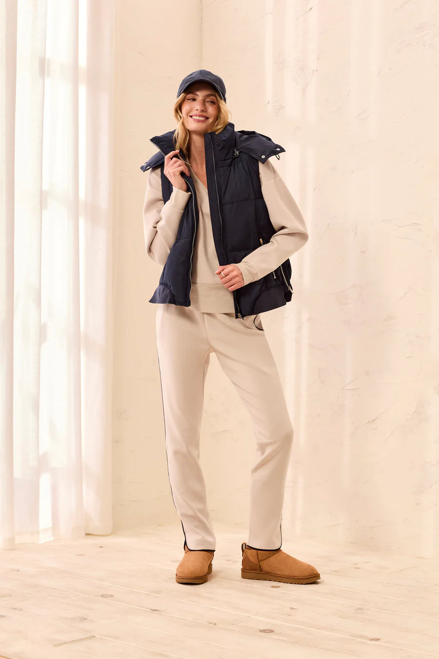 A woman modeling a dark navy puffer vest over a beige jacket and matching beige pants, styled with tan boots and a navy cap in a bright, neutral indoor setting.