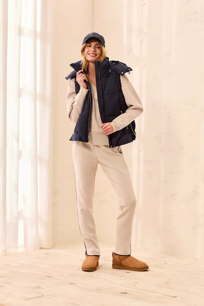 A woman modeling a dark navy puffer vest over a beige jacket and matching beige pants, styled with tan boots and a navy cap in a bright, neutral indoor setting.