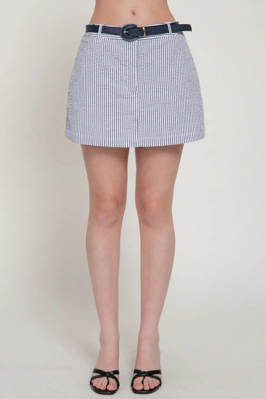 A model wearing a high-waisted, short A-line mini skirt with vertical navy and white stripes, paired with a navy blue belt, black sandals, and bare feet visible at the ankles, shot against a plain light beige studio background.