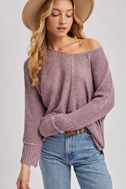 A woman wearing a chunky knit off-the-shoulder sweater in dusty mauve, paired with blue jeans and a wide-brim tan hat, against a light gray studio background.