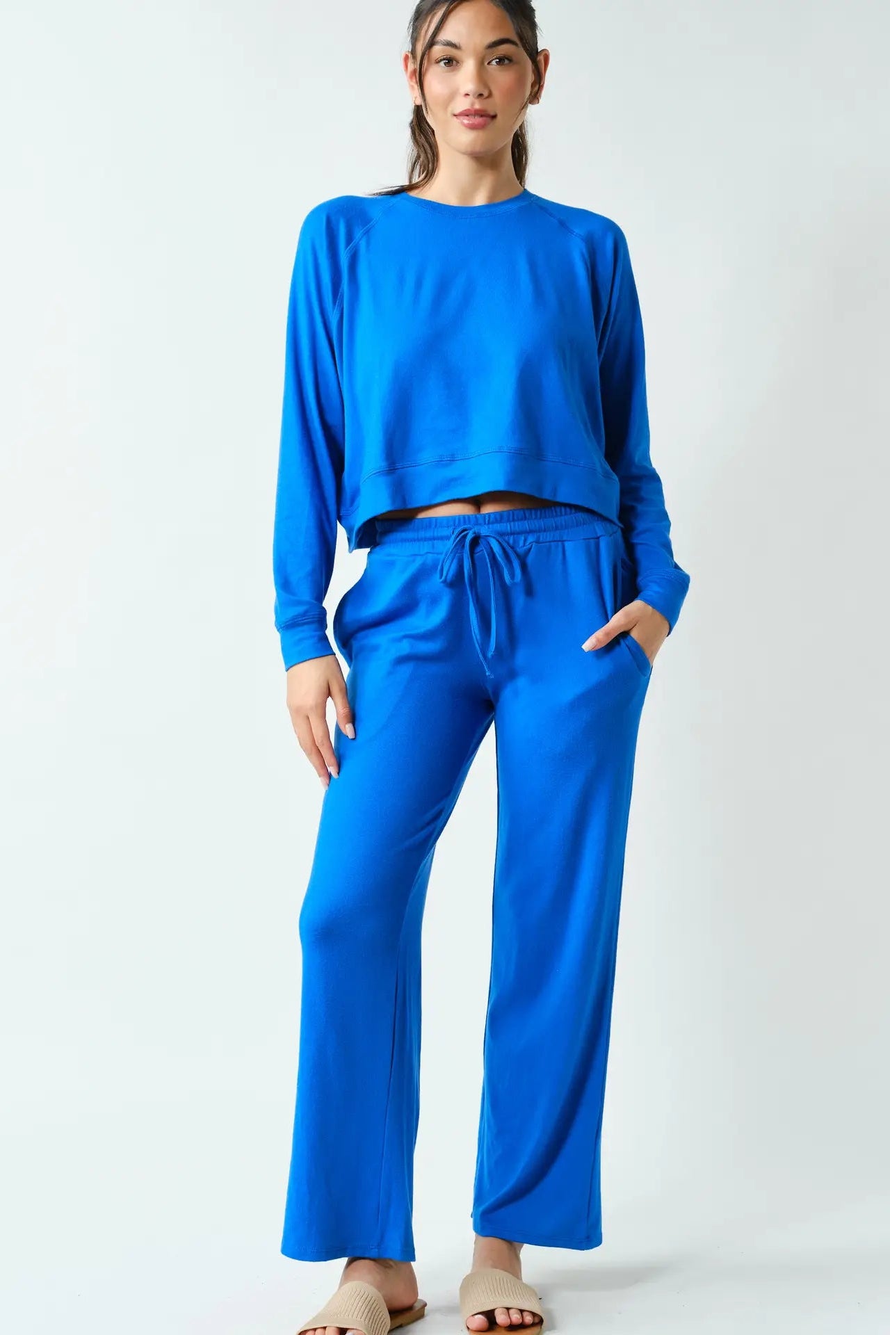 A woman modeling a bright blue two-piece loungewear set consisting of a cropped long-sleeve top and matching wide-leg drawstring pants, photographed against a clean white studio background.