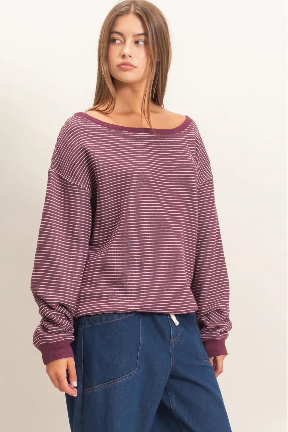 A woman modeling a long-sleeve knit top with thin horizontal stripes in dusty rose and dark red, paired with dark blue jeans, against a plain off-white background.