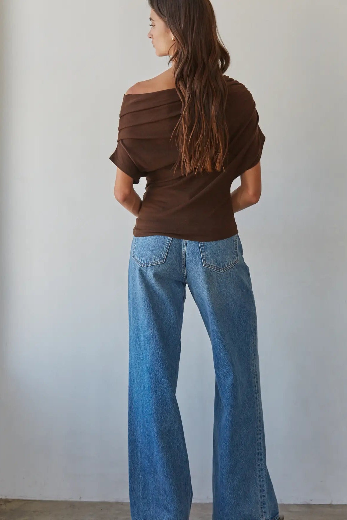 A woman shown from the back wearing a brown off-the-shoulder short-sleeve top and wide-leg blue jeans, standing against a plain off-white wall.