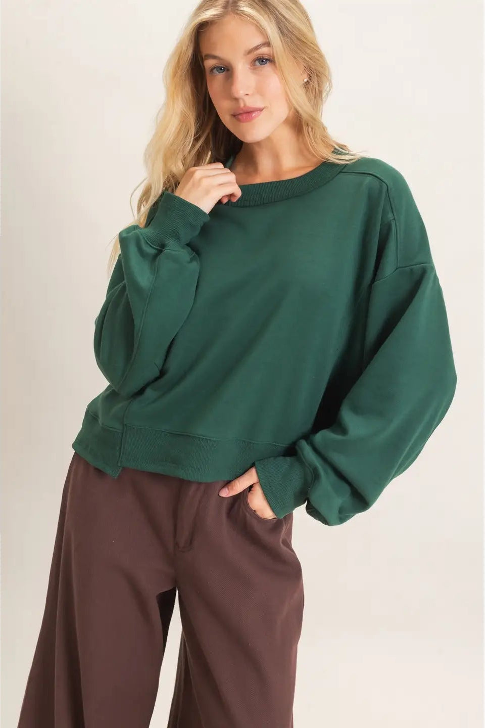 A woman wearing an oversized dark green crewneck sweatshirt and brown wide-leg pants, standing against a plain light beige studio background.