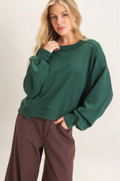 A woman wearing an oversized dark green crewneck sweatshirt and brown wide-leg pants, standing against a plain light beige studio background.