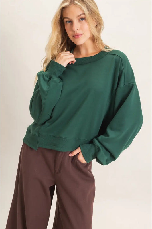 A woman wearing an oversized dark green crewneck sweatshirt and brown wide-leg pants, standing against a plain light beige studio background.