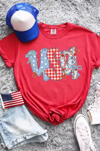 Red t-shirt with 'USA' design, blue cap, American flag, denim shorts, and pink sneakers on a gray surface.