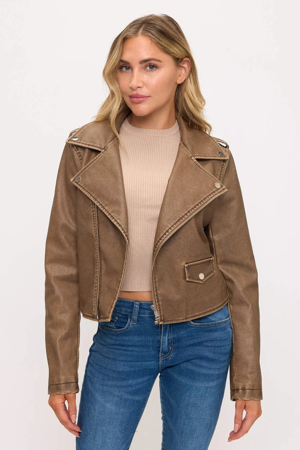 A woman modeling a cropped brown faux-leather moto jacket with silver-toned snap details, worn over a beige ribbed crop top and blue jeans, against a plain white studio background.