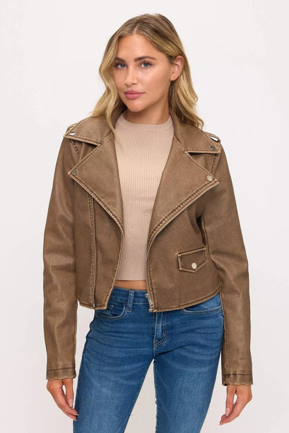 A woman modeling a cropped brown faux-leather moto jacket with silver-toned snap details, worn over a beige ribbed crop top and blue jeans, against a plain white studio background.