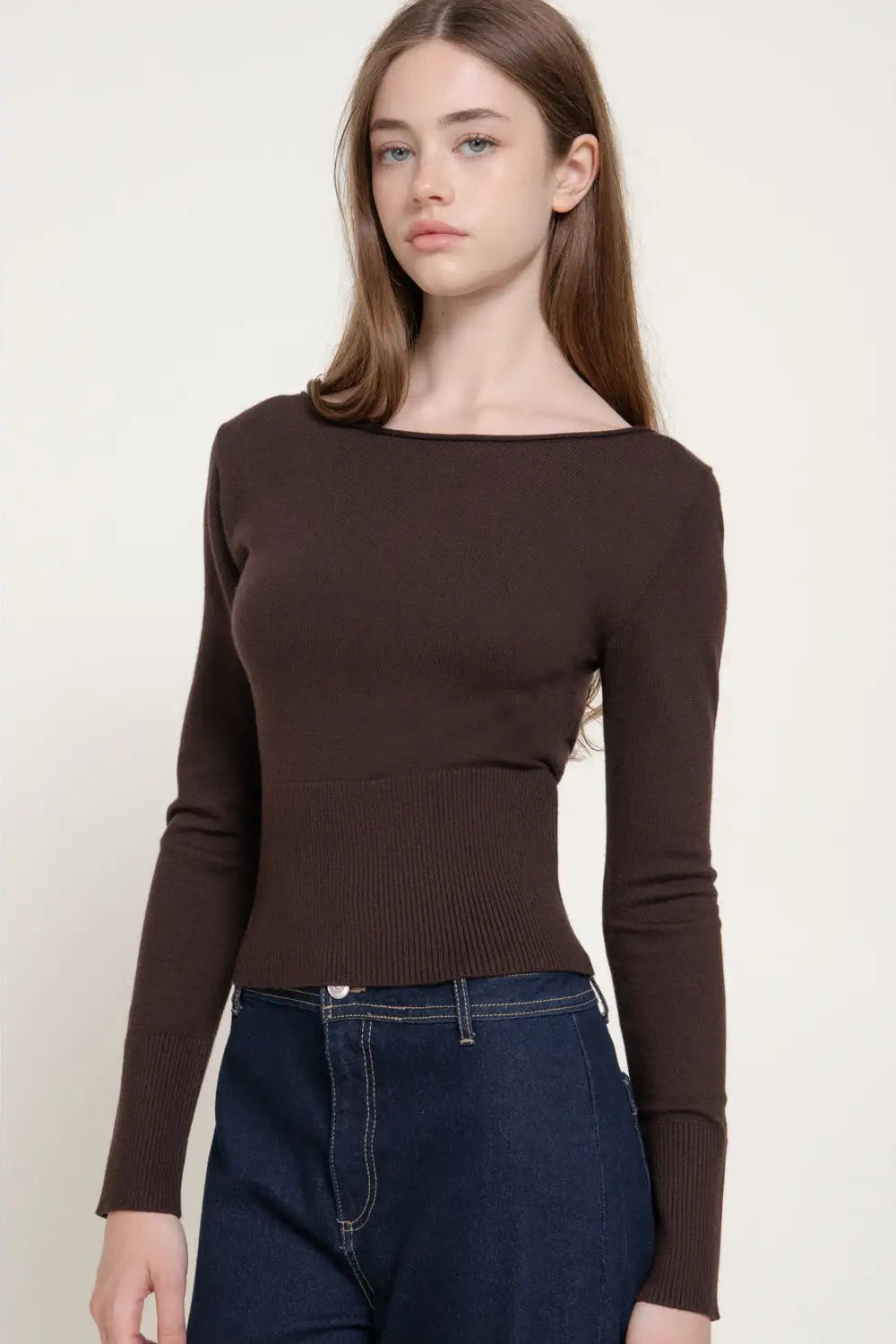 A woman modeling a dark brown long-sleeve knit top with a wide boat neckline, paired with dark blue jeans, against a plain off-white background.