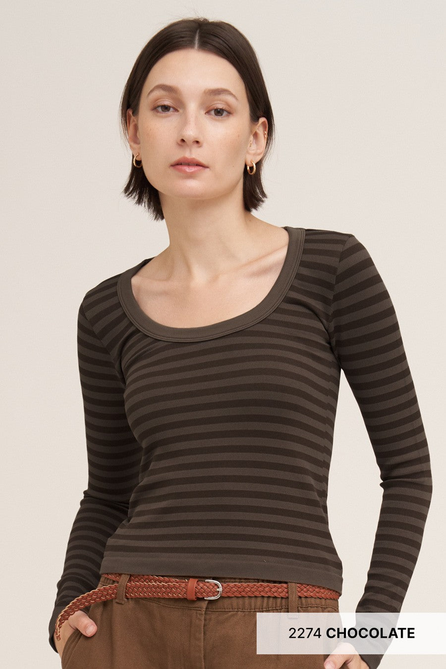 A woman modeling a dark brown and black horizontally striped long-sleeve scoop-neck top, paired with brown pants and a braided belt, against a light beige studio background.