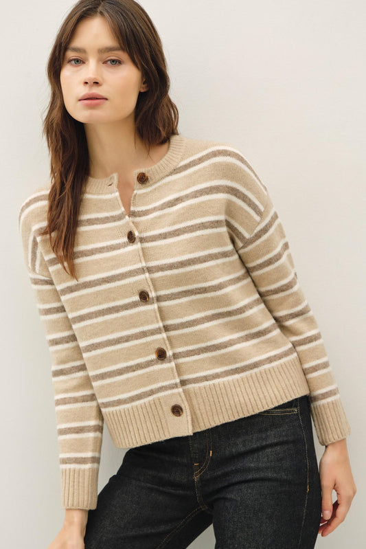 A woman wearing a beige and brown striped knit cardigan sweater with dark buttons, paired with dark jeans, against a plain light gray background.