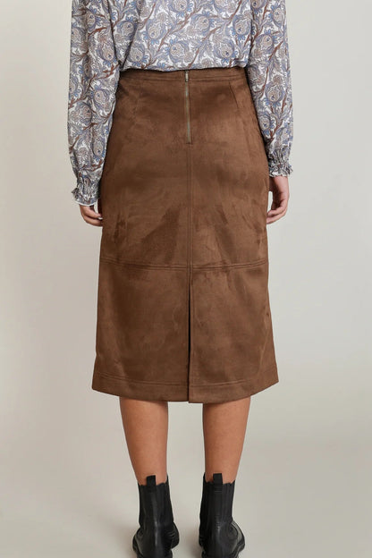 A model is shown from the back wearing a brown suede midi skirt, a patterned long-sleeve blouse, and black ankle boots against a neutral studio background.