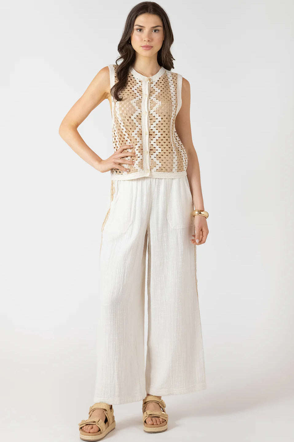 Woman wearing a sleeveless beige top with a mesh design and white pants on a white background