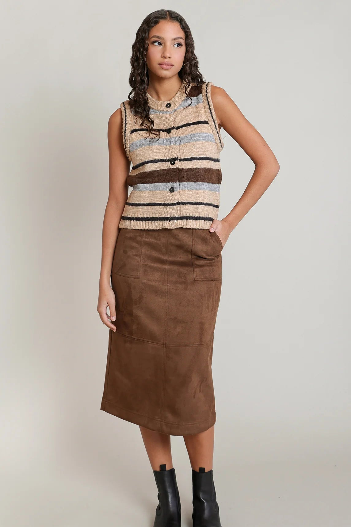 A woman modeling a sleeveless knit vest with horizontal stripes in beige, brown, and light blue, paired with a brown midi skirt and black ankle boots, against a light gray studio background.