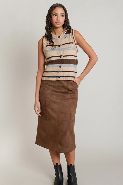 A woman modeling a sleeveless knit vest with horizontal stripes in beige, brown, and light blue, paired with a brown midi skirt and black ankle boots, against a light gray studio background.