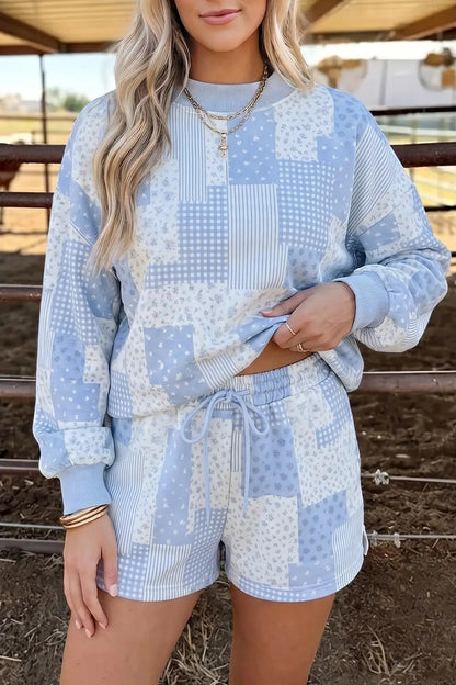 A woman wearing a light blue and white patchwork-patterned sweatshirt and matching shorts, standing in an outdoor barn setting.
