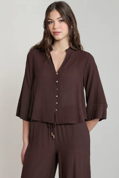 A woman modeling a dark brown button-front top with three-quarter sleeves and matching wide-leg pants against a plain light gray studio background.